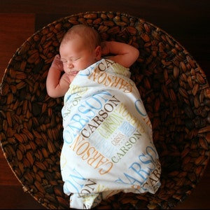 Puede incluir: Un beb&eacute; durmiendo en una cesta tejida, envuelto en una manta blanca con el nombre "Carson" impreso en azul, marr&oacute;n y amarillo.