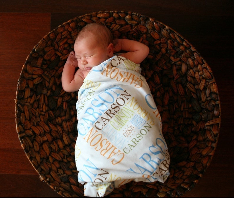 Puede incluir: Un beb&eacute; durmiendo en una cesta tejida, envuelto en una manta blanca con el nombre "Carson" impreso en azul, marr&oacute;n y amarillo.