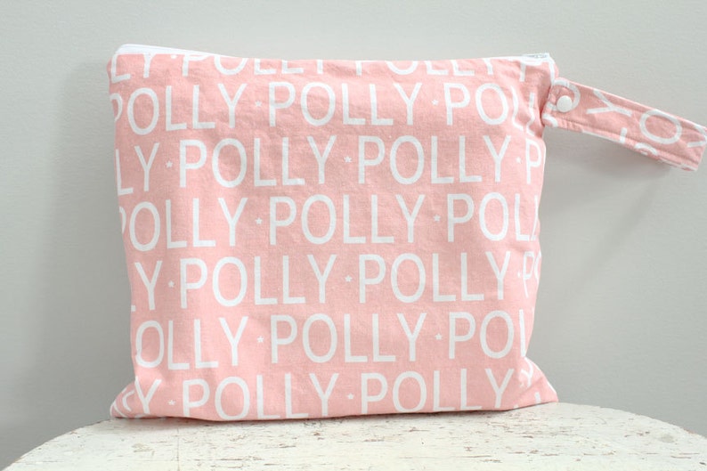 May include: Pink fabric wet bag with a white zipper and a white handle. The bag has a repeating pattern of the word 
