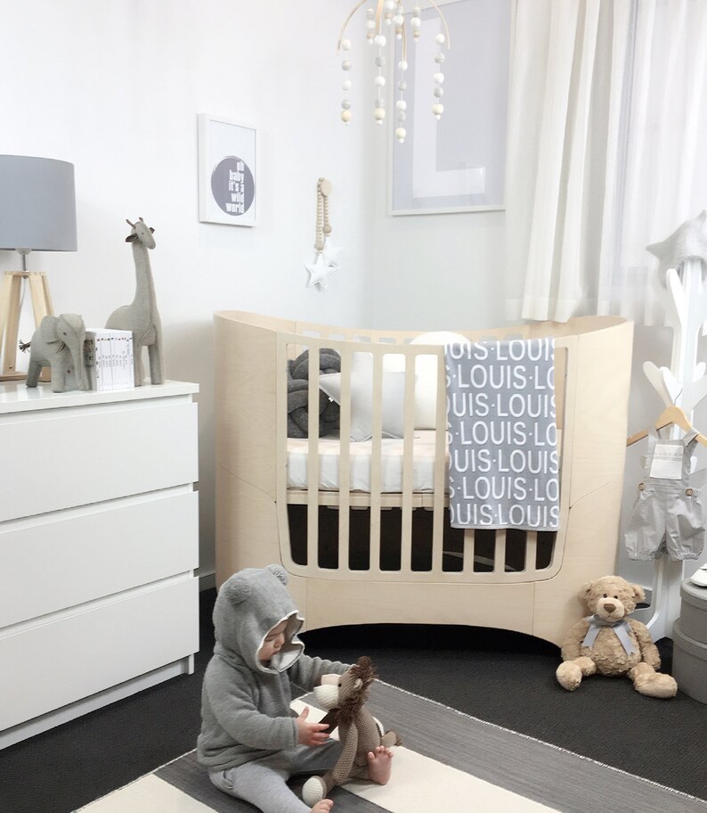 May include: A white crib with a gray and white blanket and a gray stuffed animal. The crib is in a nursery with a white dresser, a gray lamp, and a white wall with a gray and white print.