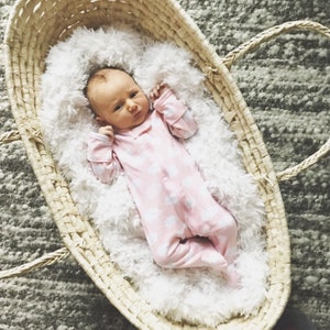 May include: A newborn baby wearing a pink and white cloud print onesie is lying in a woven wicker basket lined with a white fluffy blanket.