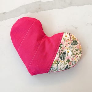 Peut inclure: Un coussin en forme de c&oelig;ur, rose vif sur une moiti&eacute; et orn&eacute; d'un motif floral sur l'autre. Le coussin est en tissu et pr&eacute;sente des coutures blanches. Le motif floral est rose et vert.