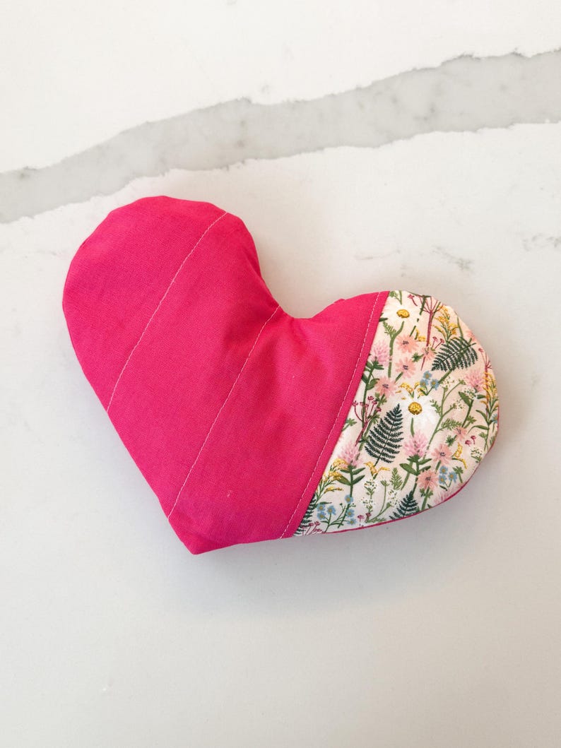 Peut inclure: Un coussin en forme de c&oelig;ur, rose vif sur une moiti&eacute; et orn&eacute; d'un motif floral sur l'autre. Le coussin est en tissu et pr&eacute;sente des coutures blanches. Le motif floral est rose et vert.