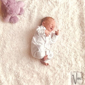 May include: A baby wearing a white onesie with small pink polka dots is lying on a white fluffy blanket. The baby is looking up and has a slightly distressed expression. A purple plush toy is in the top left corner of the image. The text "Vera" is visible in the bottom right corner of the image.