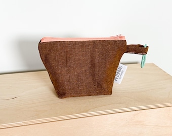 May include: A small brown zippered pouch with a pink zipper and a green clip attached to the handle. The pouch has a white label that says "PETUNIAS" and "Everyday".