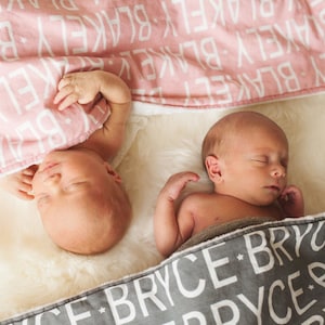 Puede incluir: Dos beb&eacute;s durmiendo en mantas personalizadas. Una manta es rosa con letras blancas que deletrean "BLAKE" y la otra es gris con letras blancas que deletrean "BRYCE".