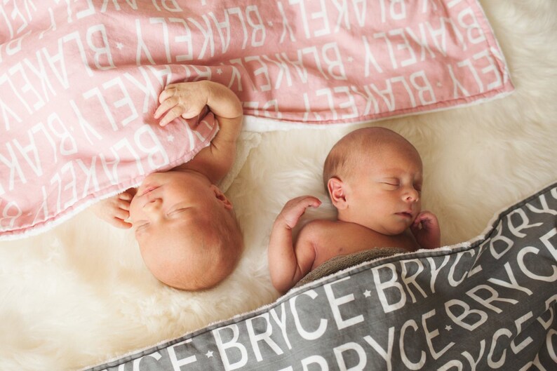 Puede incluir: Dos beb&eacute;s durmiendo en mantas personalizadas. Una manta es rosa con letras blancas que deletrean "BLAKE" y la otra es gris con letras blancas que deletrean "BRYCE".