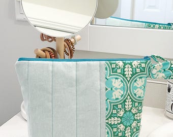 May include: A light blue quilted cosmetic bag with a teal zipper and a patterned side panel. The patterned panel features a teal and white floral and elephant design. The bag is sitting on a white countertop.