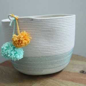 Puede incluir: Una cesta de cuerda blanca con una franja azul claro alrededor de la parte superior. La cesta tiene dos pompones unidos al asa, uno amarillo y otro azul claro.
