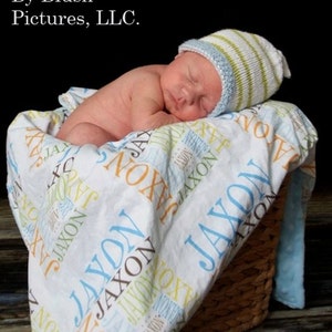 Puede incluir: Un beb&eacute; reci&eacute;n nacido duerme sobre una manta blanca con el nombre "Jaxon" impreso en ella en varios colores. El beb&eacute; lleva un gorro de punto a rayas azules y verdes.
