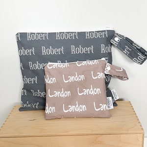 Pode incluir: Duas bolsas de zíper de tecido com alças. A bolsa maior é cinza com texto branco que diz "Robert" repetidamente. A bolsa menor é marrom com texto branco que diz "Landon" repetidamente. A bolsa menor tem uma etiqueta branca que diz "PETUNIAS by Melanie"