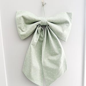 May include: A large, sage green bow with white polka dots hangs against a white wall. The bow is made of fabric and has long, flowing tails. The bow is tied in the center and suspended by a clear hook.