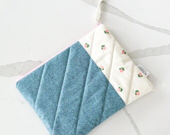May include: A quilted zippered pouch with a blue denim fabric on one side and a white fabric with a pink peach pattern on the other side. The pouch has a pink zipper and a white pull tab.