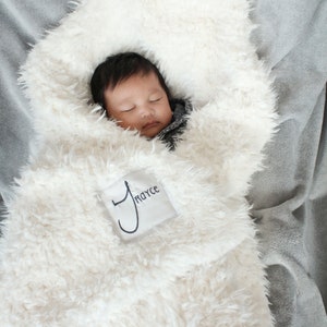 May include: A white faux fur baby blanket with a personalised embroidered name tag that says "Royce". A baby is sleeping peacefully inside the blanket.