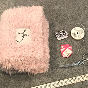 May include: A pink fuzzy blanket with the name "Jama" embroidered on it. The blanket is surrounded by craft supplies, including a spool of white ribbon, a pair of scissors, and small gift tags.