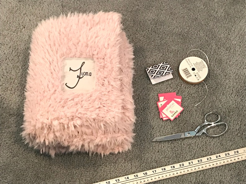 May include: A pink fuzzy blanket with the name "Jama" embroidered on it. The blanket is surrounded by craft supplies, including a spool of white ribbon, a pair of scissors, and small gift tags.
