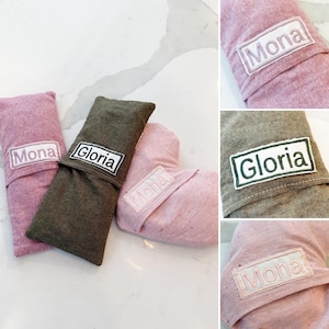 Peut inclure: Trois articles en tissu dans les tons de rose et vert olive, chacun avec un patch rectangulaire blanc brod&eacute; d'un nom. Les noms "Mona" et "Gloria" sont visibles. Les articles semblent &ecirc;tre de petits oreillers ou pochettes.