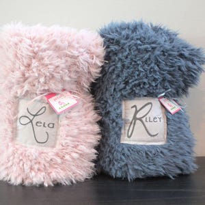 May include: Two personalised fluffy blankets, one pink with the name "Ela" and one grey with the name "Riley".