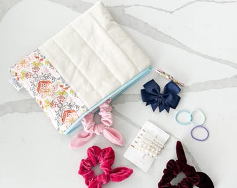 May include: A white quilted zippered pouch with a floral print on the bottom half. The pouch is open and contains a variety of hair accessories, including pink, red, and burgundy scrunchies, a navy blue bow, and a set of pearl hair clips.