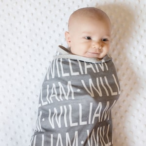 May include: A baby swaddled in a gray and white personalized blanket with the name "William" repeated in a pattern.