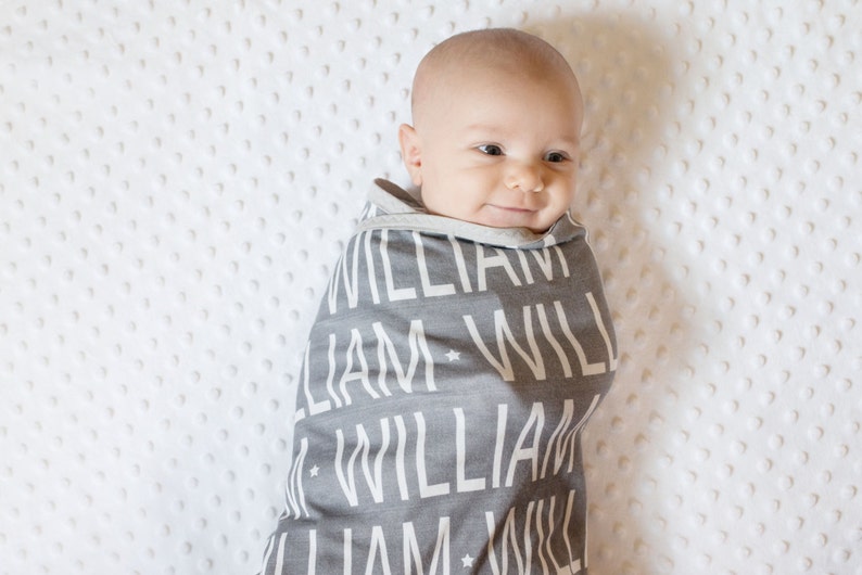 May include: A baby swaddled in a gray and white personalized blanket with the name "William" repeated in a pattern.