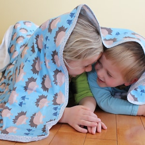 May include: A blue hooded baby blanket with a pattern of brown hedgehogs and blue mushrooms. The blanket has a white interior and a light blue trim. Two children are under the blanket, smiling and looking at each other.