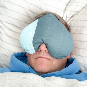 Peut inclure: Un masque pour les yeux en forme de c&oelig;ur, bleu et blanc, pos&eacute; sur le visage d'une personne. Le masque est en tissu, avec une section bleu clair &agrave; pois blancs. La personne porte un sweat &agrave; capuche bleu et est couverte d'une couverture blanche.