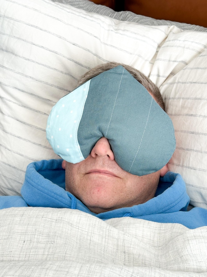 Peut inclure: Un masque pour les yeux en forme de c&oelig;ur, bleu et blanc, pos&eacute; sur le visage d'une personne. Le masque est en tissu, avec une section bleu clair &agrave; pois blancs. La personne porte un sweat &agrave; capuche bleu et est couverte d'une couverture blanche.