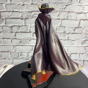 K&ouml;nnte beinhalten: Figur einer Figur mit einem dunkelvioletten Umhang mit goldenen Verzierungen. Die Figur steht auf einem schwarzen Sockel mit roten und goldenen Akzenten. Der Hintergrund ist eine wei&szlig;e Backsteinmauer.
