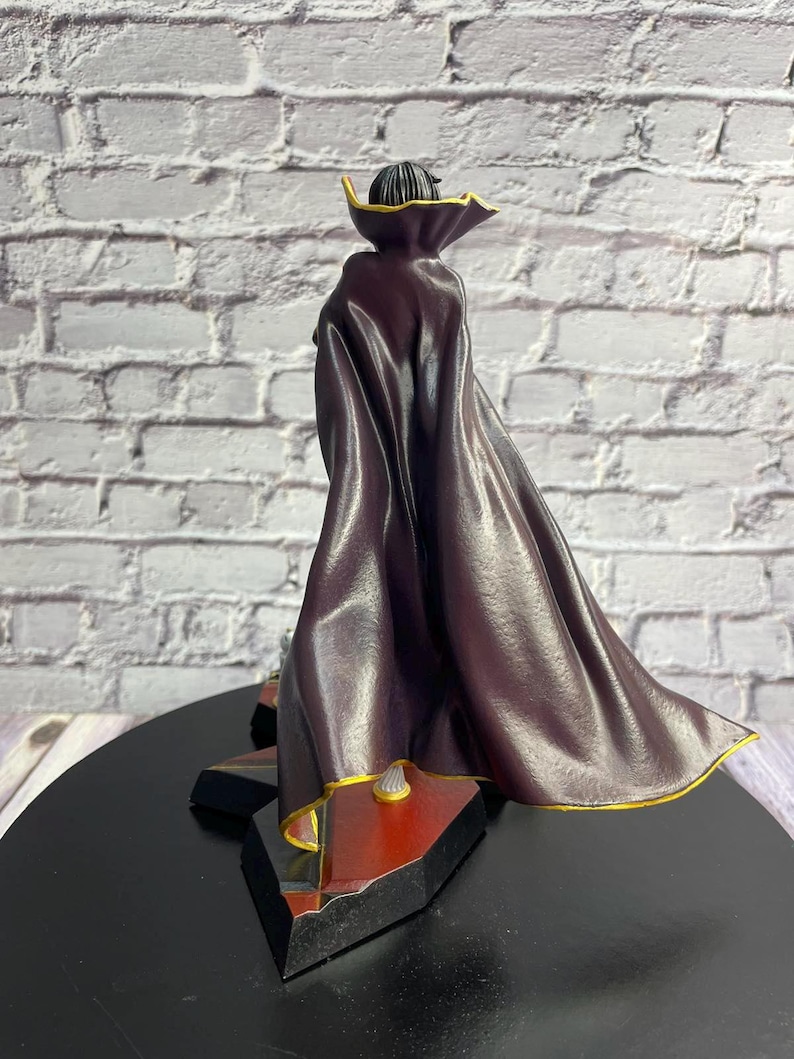 K&ouml;nnte beinhalten: Figur einer Figur mit einem dunkelvioletten Umhang mit goldenen Verzierungen. Die Figur steht auf einem schwarzen Sockel mit roten und goldenen Akzenten. Der Hintergrund ist eine wei&szlig;e Backsteinmauer.