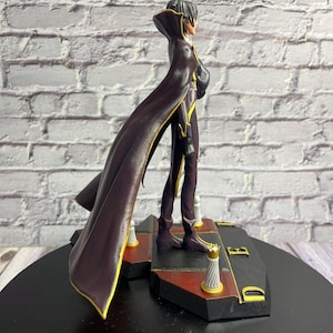 K&ouml;nnte beinhalten: Detaillierte Figur einer Figur in einem langen, dunkelvioletten Mantel mit goldenen Verzierungen. Die Figur steht auf einem schwarz-roten Sockel mit goldenen Akzenten. Die Figur hat schwarzes Haar und ist im Seitenprofil dargestellt.