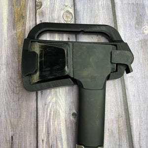 Peut inclure: Un étui de protection en plastique noir recouvrant la tête métallique d'une hachette Fiskars. L'étui présente une boucle de poignée rectangulaire sur le dessus et un mécanisme de verrouillage sur le côté. La hachette a un manche en bois.