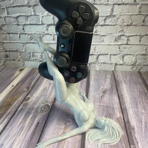 May include: A black PlayStation 4 controller held by a light gray female figure. The figure is in a reclining pose, with long flowing hair. The controller is the focal point, with its buttons and joysticks visible. The background is a wooden surface.