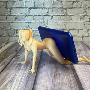 May include: A nude-colored figurine in a crawling pose, supporting a blue smartphone. The figurine has blonde pigtails and is positioned on a wooden surface. The phone is held in place by the figurine's back.