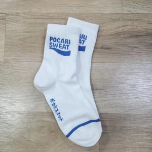 Może przedstawiać: Para białych skarpet do kostek z niebieskim wykończeniem. Jedna skarpeta ma napis "Pocari Sweat" w kolorze niebieskim z niebieskim wzorem fali. Druga skarpeta ma napis "Pocari Sweat" w języku japońskim.