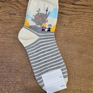 May include: A pair of white socks with a gray and white striped pattern on the foot and a cartoon graphic of a castle and two figures on the upper portion. A small white tag is attached to the sock.
