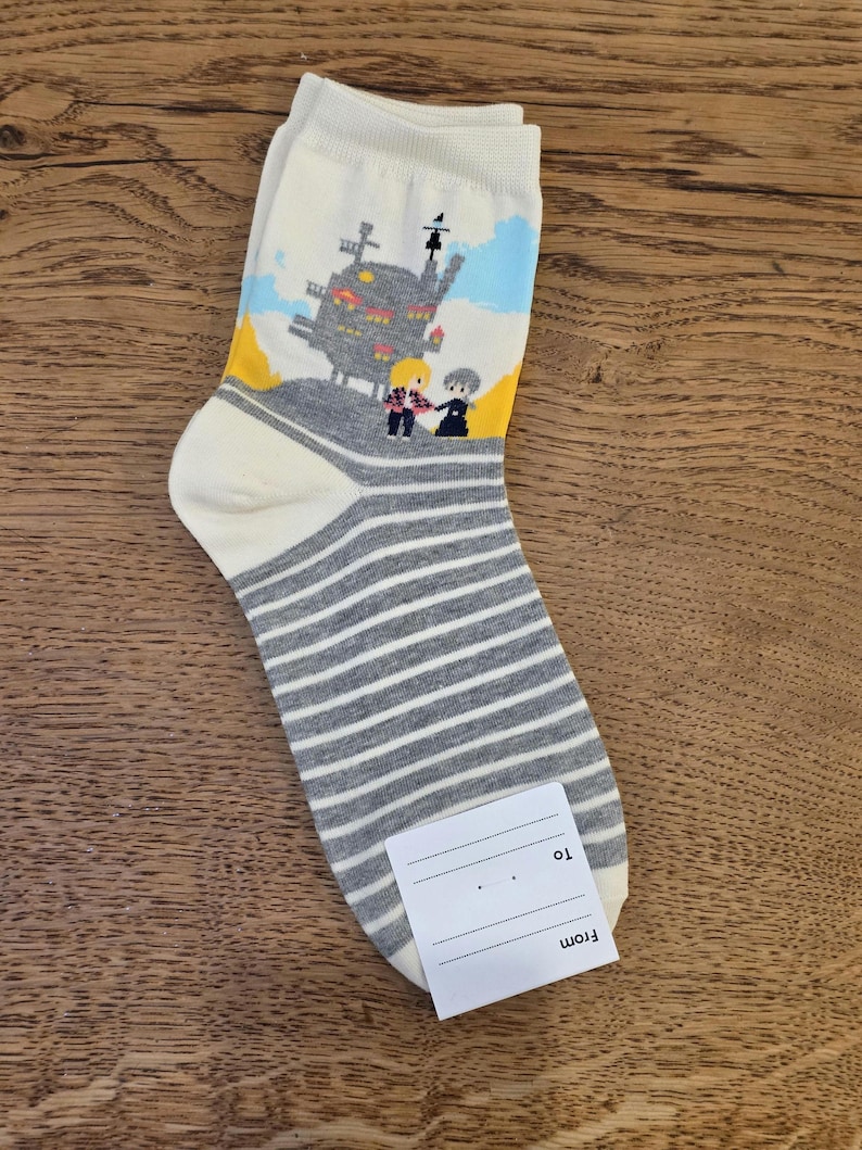 May include: A pair of white socks with a gray and white striped pattern on the foot and a cartoon graphic of a castle and two figures on the upper portion. A small white tag is attached to the sock.
