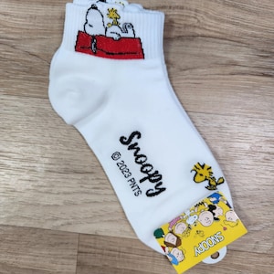 以下が含まれることがあります： Snoopyが犬小屋で寝ている漫画の画像が描かれた白い足首丈の靴下。靴下には「Snoopy」と「© 2023 PNTS」の文字が印刷されています。靴下は、「Snoopy」と黒文字で書かれた黄色のタグで包装されています。