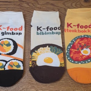 Peut inclure: Trois paires de chaussettes avec des motifs de plats coréens. Une paire représente du gimbap, une autre du bibimbap et la troisième du tteokbokki. Chaque chaussette porte l'inscription "K-food" au-dessus de l'image du plat. Les chaussettes sont blanches, beiges et jaunes.