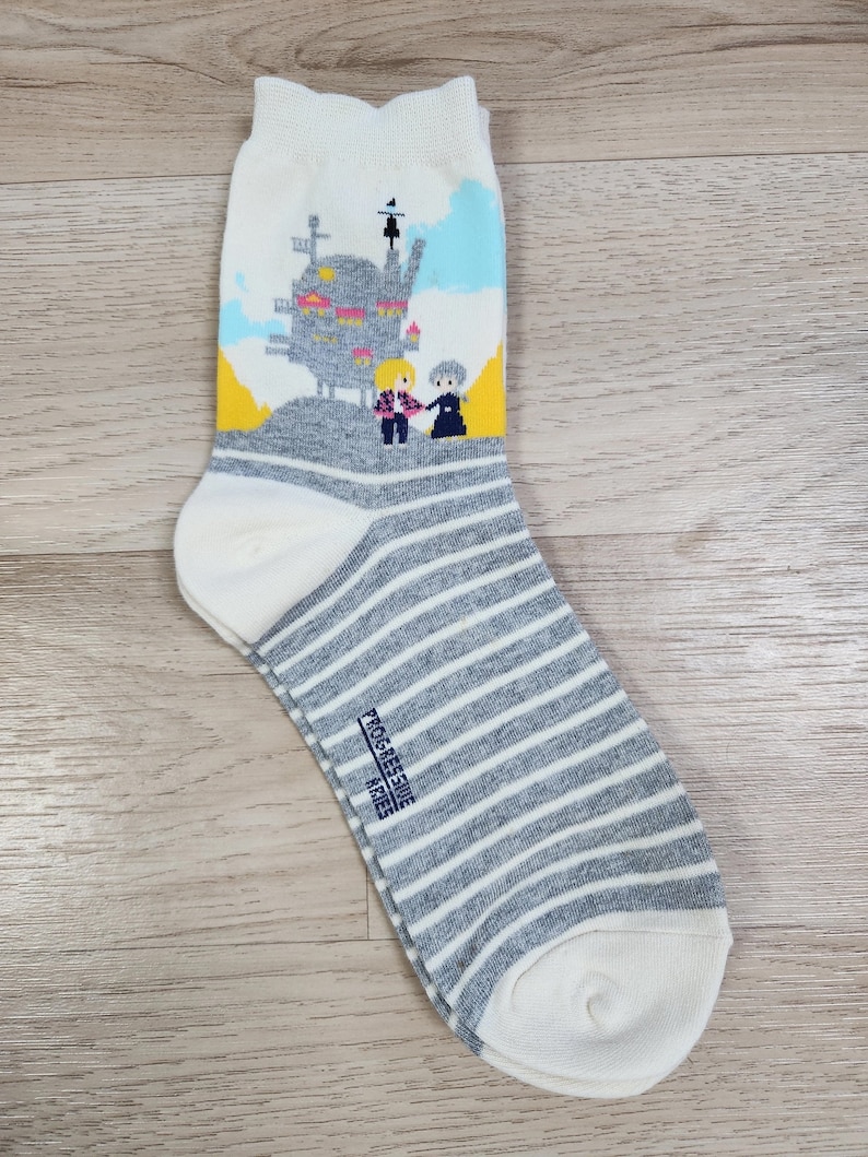 K&ouml;nnte beinhalten: Wei&szlig;e Socken mit einem grau-wei&szlig;en Streifenmuster. Die Socken zeigen ein Motiv mit einem fliegenden Haus, vor dem zwei Figuren stehen. Der Text "PROGRESSIVE ARIES" ist auf den Socken aufgedruckt.