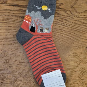May include: Gray and orange socks with a Japanese anime-style design. The top of the sock features a scene with a building, characters, and a sun. The lower portion has orange and gray horizontal stripes. A white tag is attached.