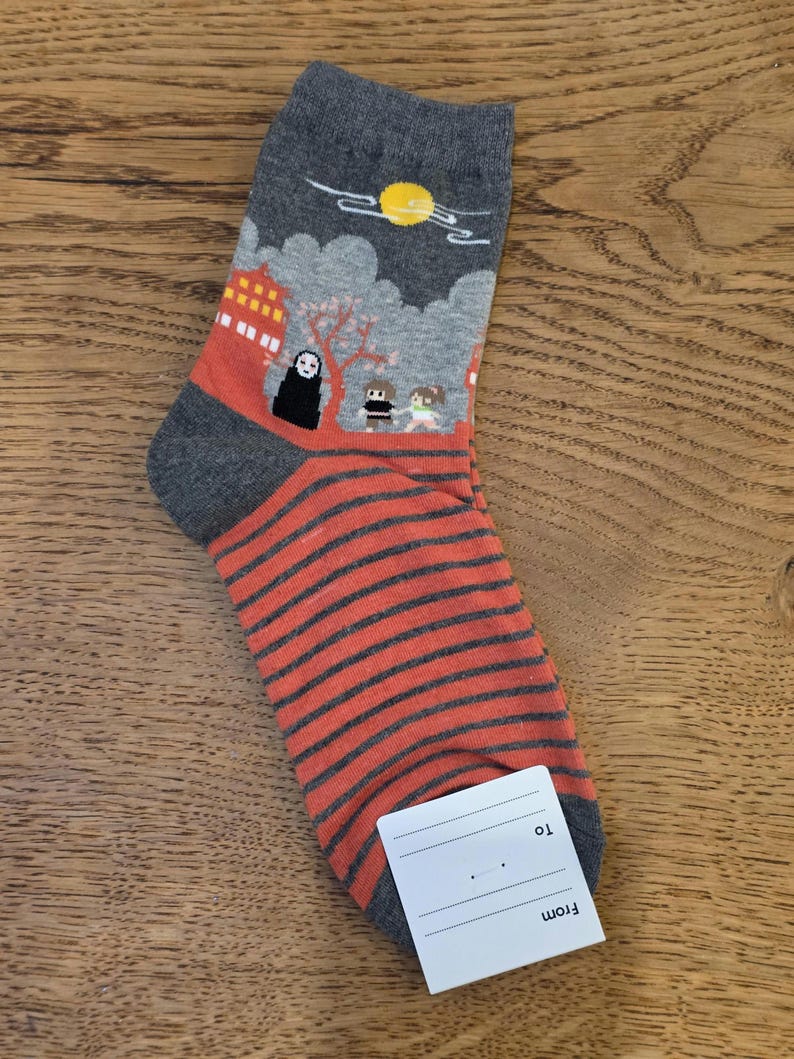 May include: Gray and orange socks with a Japanese anime-style design. The top of the sock features a scene with a building, characters, and a sun. The lower portion has orange and gray horizontal stripes. A white tag is attached.