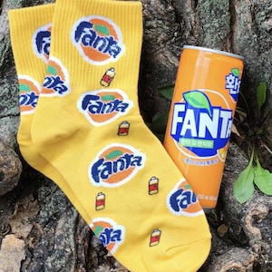 Könnte beinhalten: Gelbe Socken mit einem sich wiederholenden Muster des Fanta-Logos und einer kleinen orangenen Dose. Die Socken liegen auf einem Baumstamm mit einer Dose Fanta.