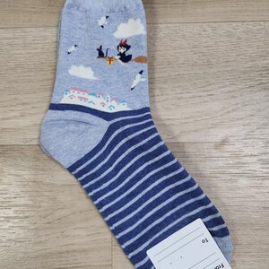 May include: A pair of grey and blue striped socks with a whimsical design featuring a witch flying on a broomstick with a black cat and a gift box. The socks have a white tag with the words "From" and "To" on it.