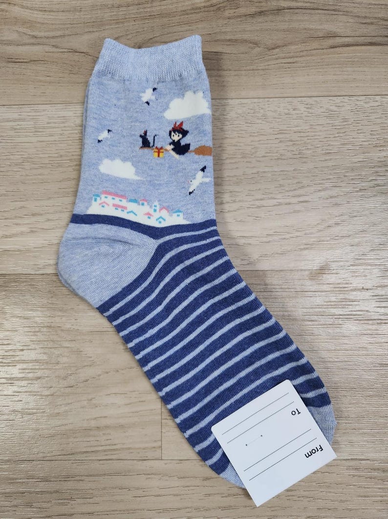 May include: A pair of grey and blue striped socks with a whimsical design featuring a witch flying on a broomstick with a black cat and a gift box. The socks have a white tag with the words "From" and "To" on it.