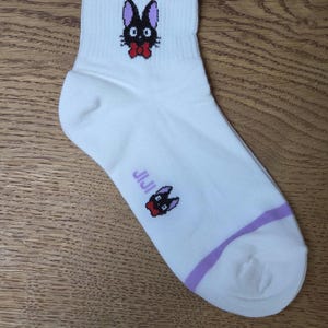 May include: White ankle sock featuring a black cat illustration with a red bow tie. The sock has a purple stripe near the ankle and the word "JIJI" printed on the side. The cat illustration is repeated on the top of the sock.