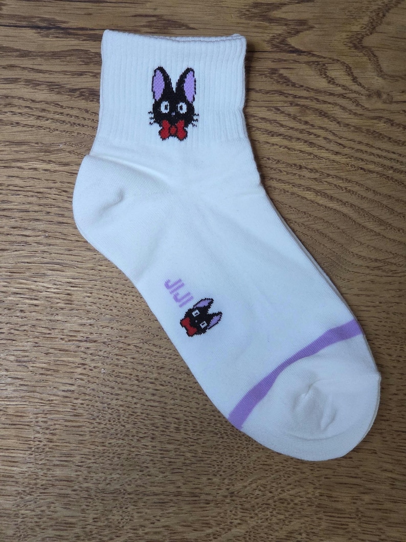 May include: White ankle sock featuring a black cat illustration with a red bow tie. The sock has a purple stripe near the ankle and the word "JIJI" printed on the side. The cat illustration is repeated on the top of the sock.