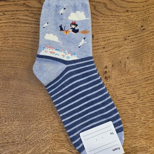 May include: A light blue sock with a witch riding a broom, clouds, and a town scene. The sock has dark blue and white horizontal stripes on the lower portion. A white tag is attached.
