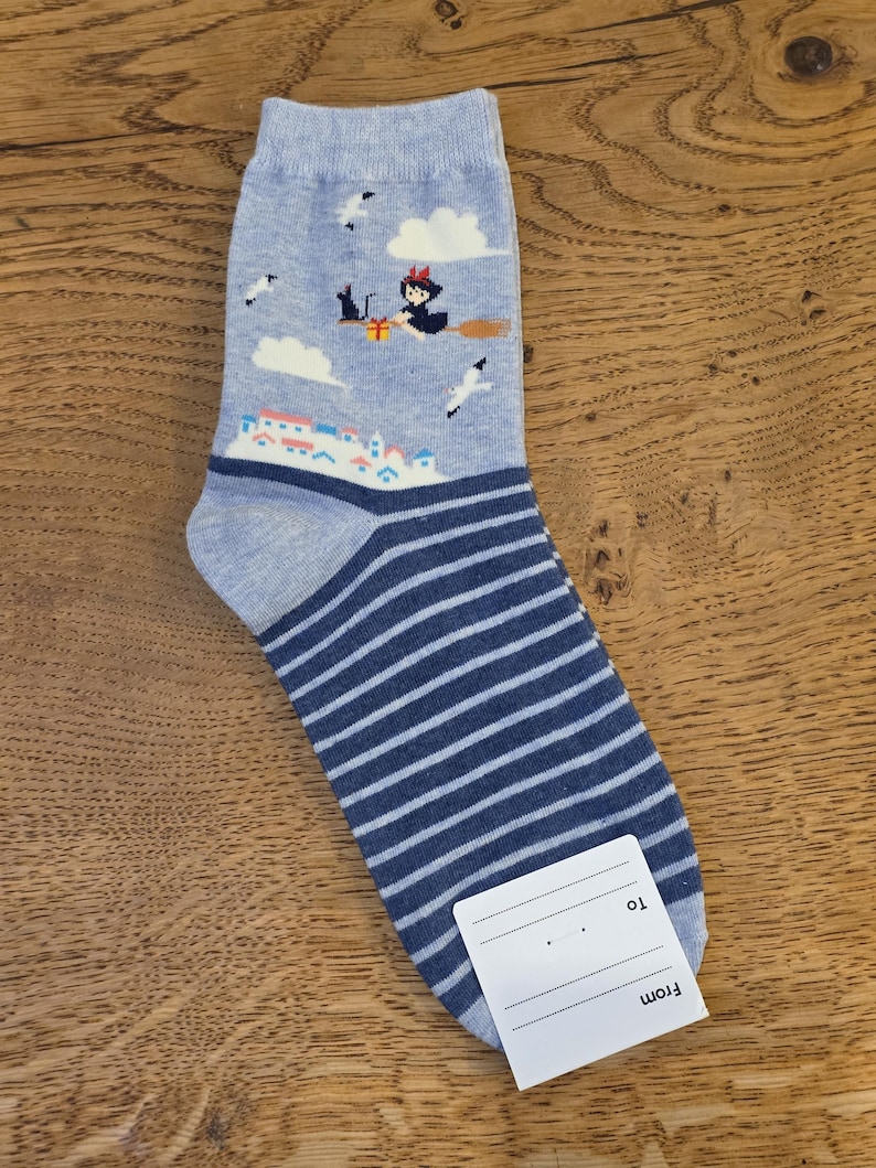 May include: A light blue sock with a witch riding a broom, clouds, and a town scene. The sock has dark blue and white horizontal stripes on the lower portion. A white tag is attached.
