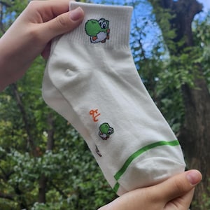 Puede incluir: Calcetines blancos de tobillo con un personaje Yoshi verde y naranja bordado. Los calcetines tienen un ribete verde alrededor de la parte superior.